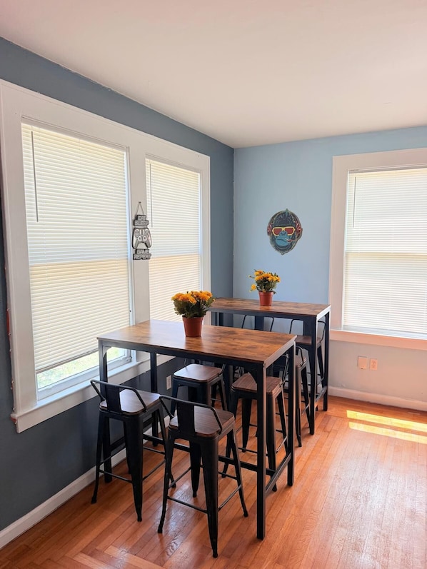 Un comedor con una mesa de madera y sillas negras, ventanas amplias con persianas y un adorno colgado en la pared.