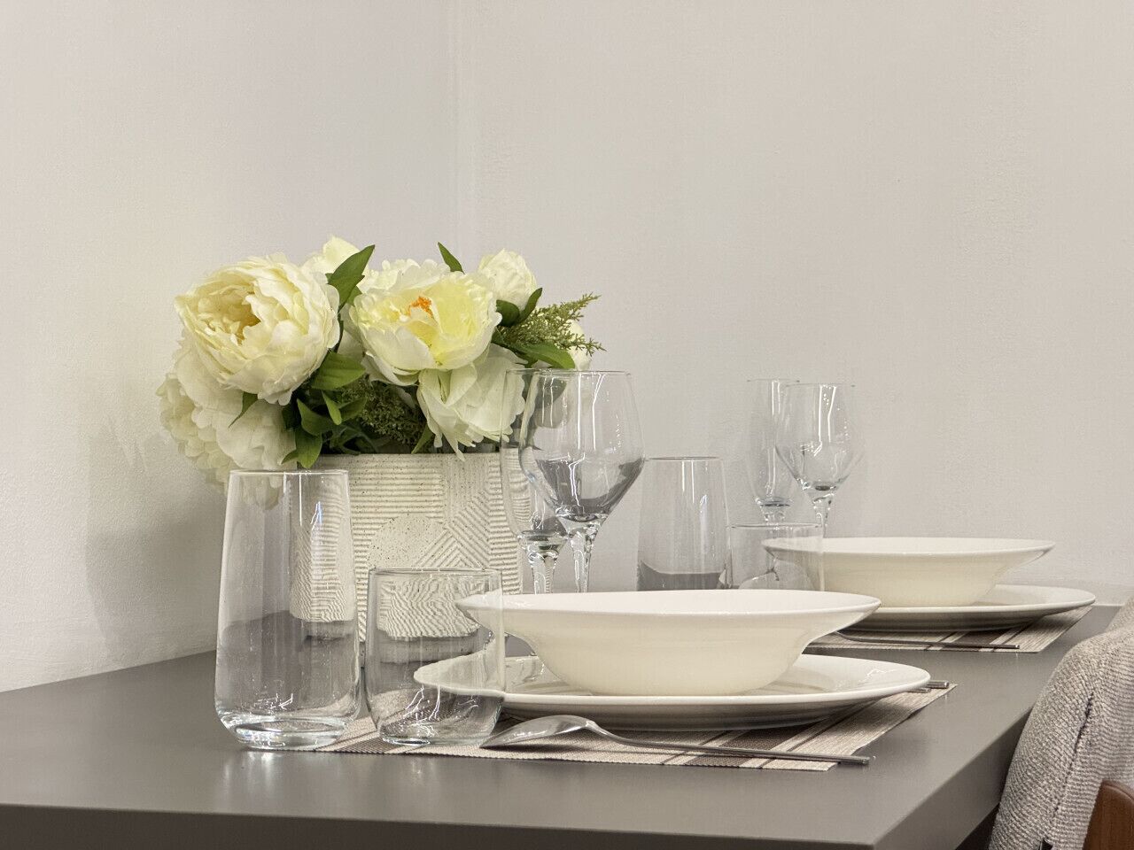 Een eettafel gedekt met witte borden, kommen en glazen, met een vaas witte bloemen.