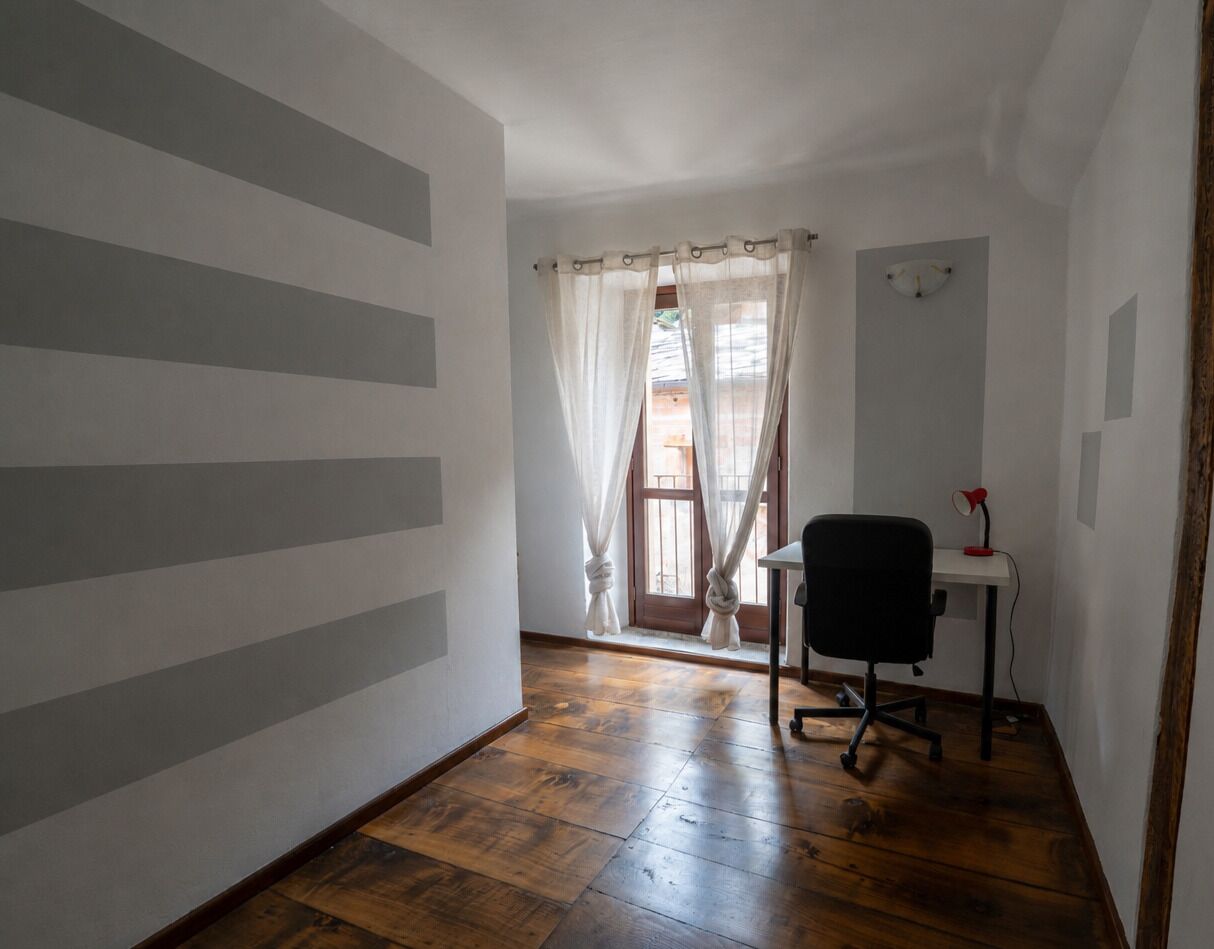 Ein Zimmer mit Holzboden, einem Schreibtisch mit Stuhl, einem Fenster mit Vorhängen und einer Wand mit Streifenmuster.