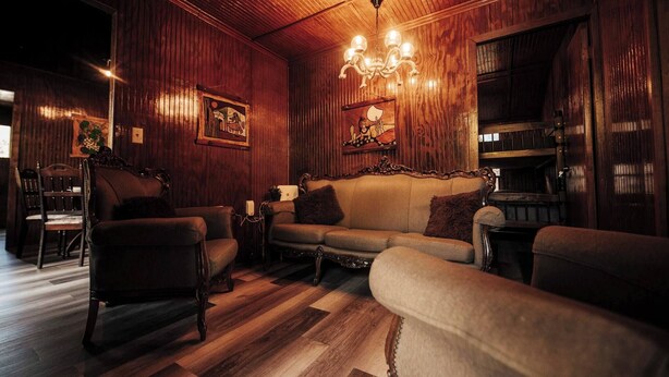A cozy room with wooden walls, a sofa, two armchairs, a chandelier, and a dining area in the background. - Naranjito River Retreat | Events & Groups | Sleeps 13 (Naranjito)