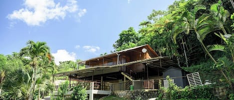 Ein zweistöckiges Holzhaus mit einem Balkon, umgeben von üppigem Grün und blauem Himmel.