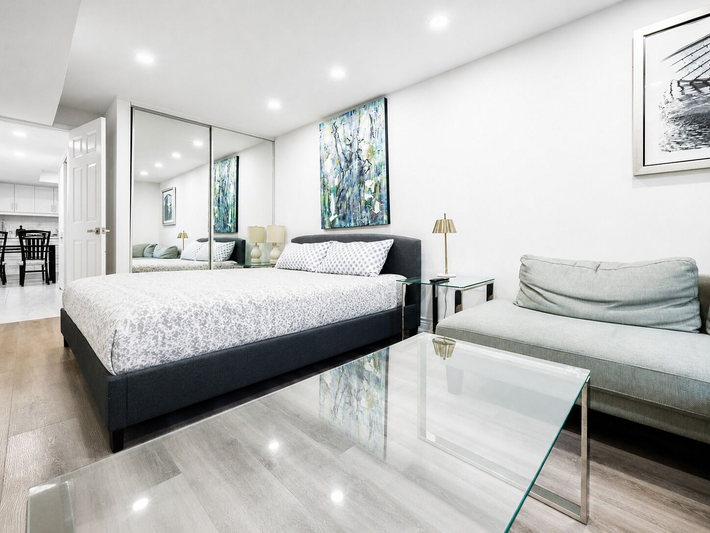 A modern bedroom with a large bed, a sofa, a glass coffee table, and artwork on the walls.