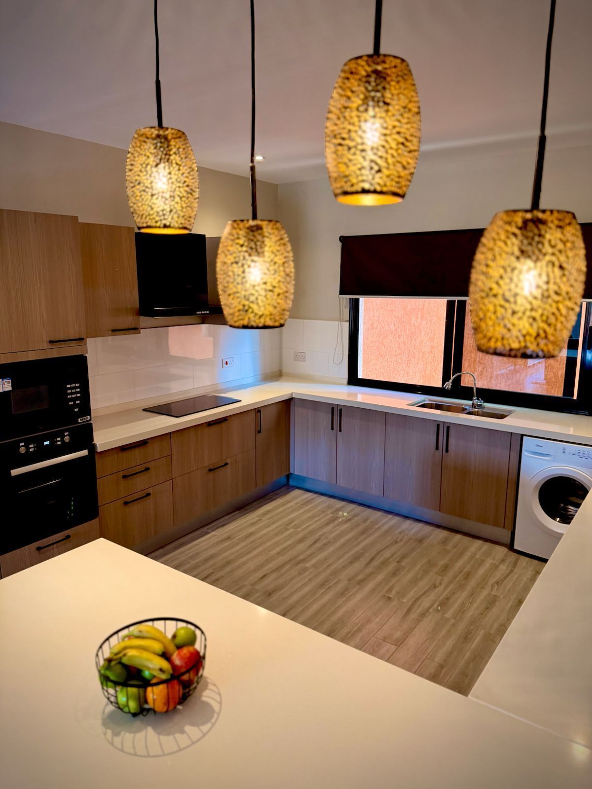 A modern kitchen with wooden cabinets, a built-in oven, a washing machine, and a fruit bowl on the counter.
