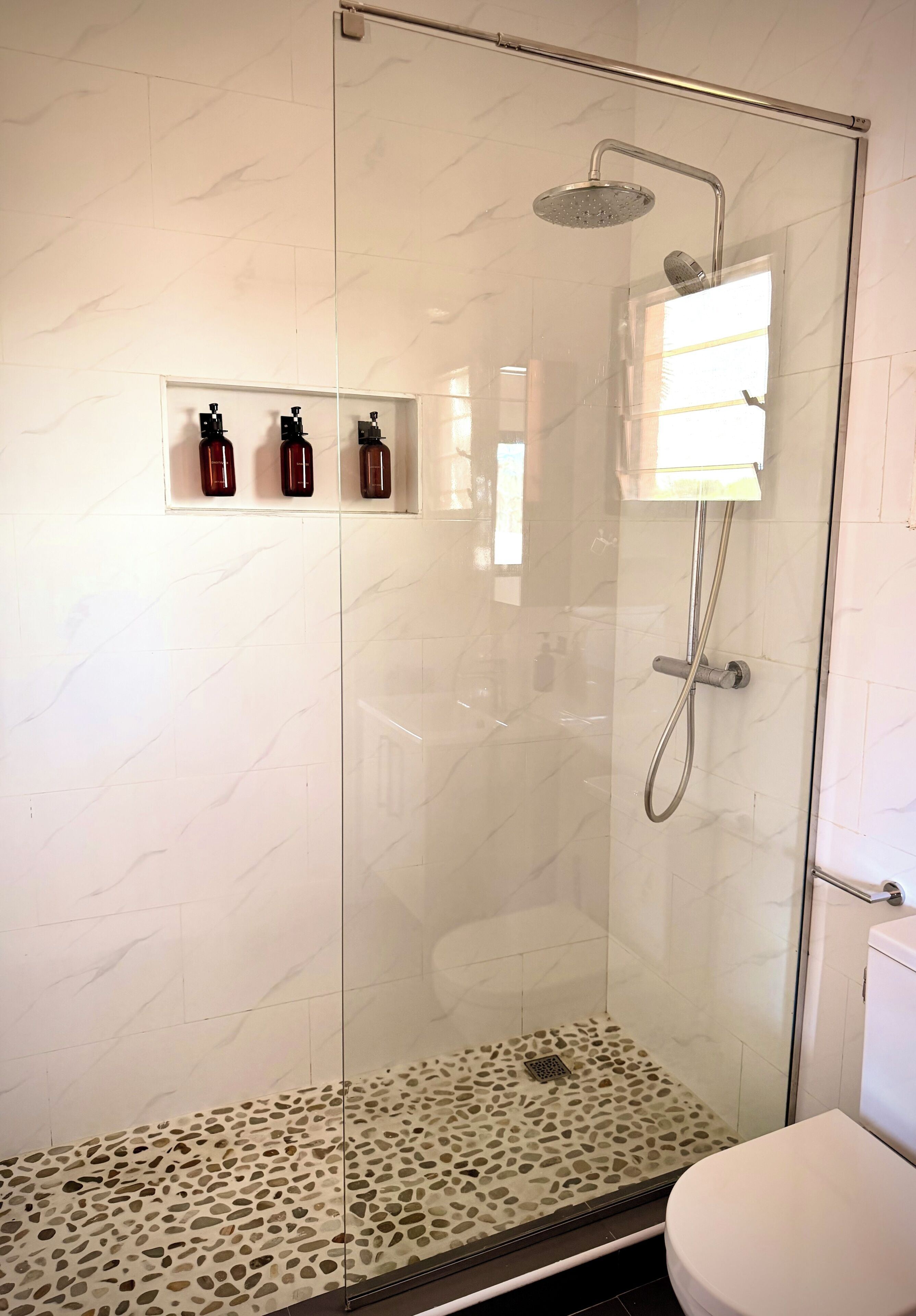 A glass-enclosed shower with a rain showerhead, a bench with a pebble-patterned floor, and three bottles on a shelf.
