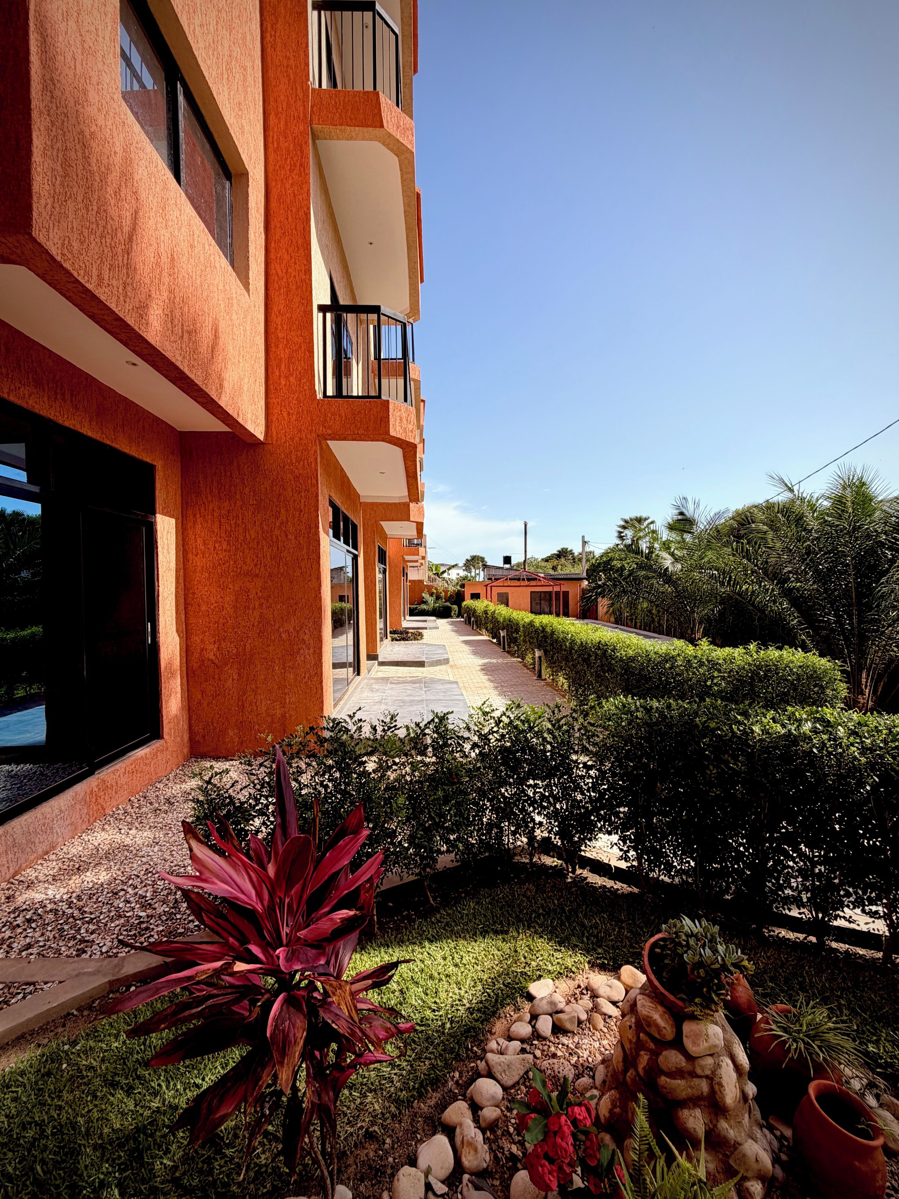 A modern building with a pathway leading to a gated entrance, surrounded by well-maintained landscaping and a clear sky.