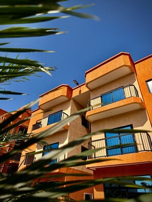 A multi-story building with balconies, a clear blue sky, and palm leaves in the foreground. - Teranga Tides (Bijilo)