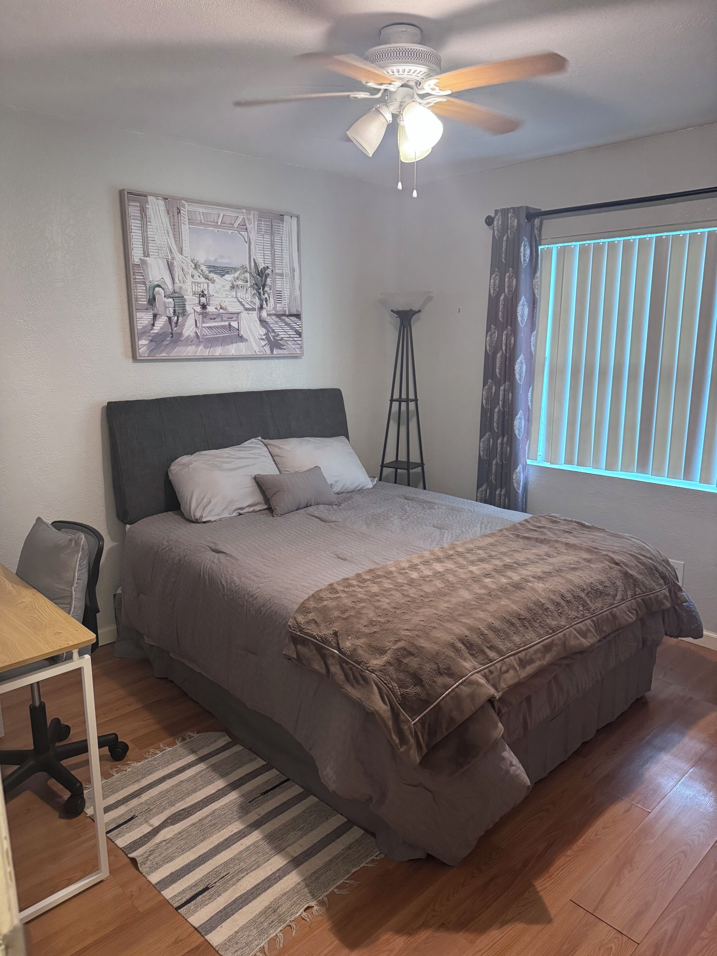 A bedroom with a bed, a desk, a standing lamp, and a ceiling fan.
