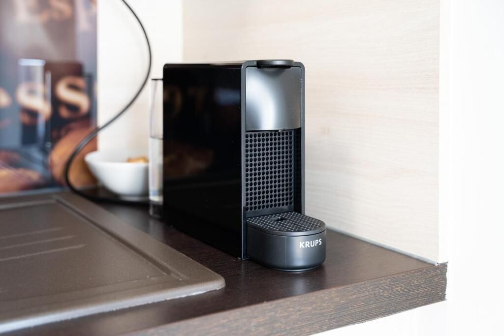 A black KRUPS speaker on a wooden surface with a laptop and a white bowl in the background.