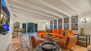A spacious living room with a large orange sofa, a coffee table, and a fireplace. There are framed artworks on the walls and a view of the outdoors through large windows. - Desert Dreams Ranch - Idyllic 6 BR Next to the Polo Grounds (Indio)