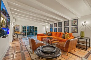 Ein geräumiges Wohnzimmer mit einem großen, orangefarbenen Sofa, einem Couchtisch und einem Kamin. An den Wänden befinden sich gerahmte Kunstwerke und durch große Fenster ist die Außenwelt zu sehen.