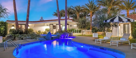 A swimming pool with a blue-lit edge, surrounded by lounge chairs and palm trees.