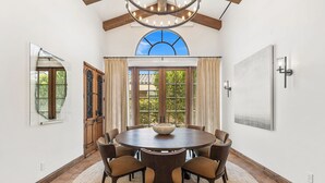 Uma sala de jantar com uma mesa redonda, seis cadeiras, um candelabro e uma janela grande com cortinas.