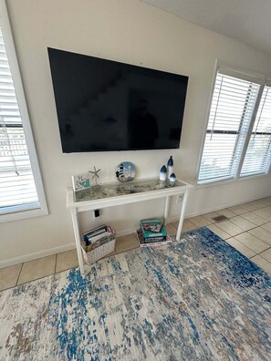 A living room with a large flat-screen TV mounted on the wall, a white console table with decorative items, and a patterned rug on the floor. - West end, great views, steps to beach, new rental! (Oak Island)