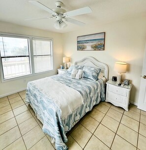Quarto com cama, duas mesas de cabeceira, um ventilador de teto, janela com persianas e um quadro na parede.