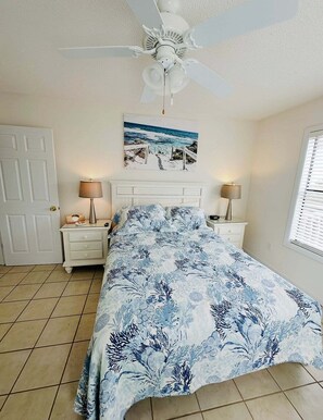 A bedroom with a bed, two bedside tables, a ceiling fan, a wall-mounted painting, a door, and a window with blinds. - West end, great views, steps to beach, new rental! (Oak Island)