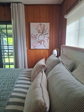 A bedroom with a bed, pillows, a painting, wooden walls, and a window.
