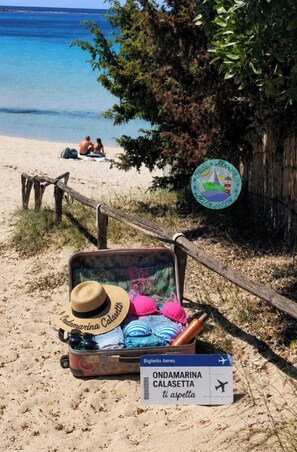 A beach scene with a suitcase containing a straw hat, sunglasses, and a pink cap, a sign with 'Biglietto Aereo' and 'ONDAMARINA CALASETTA' text, and a wooden fence with a signboard.
