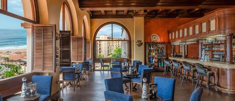 A restaurant with a view of the beach, featuring blue upholstered chairs, a bar area, and a large arched window.