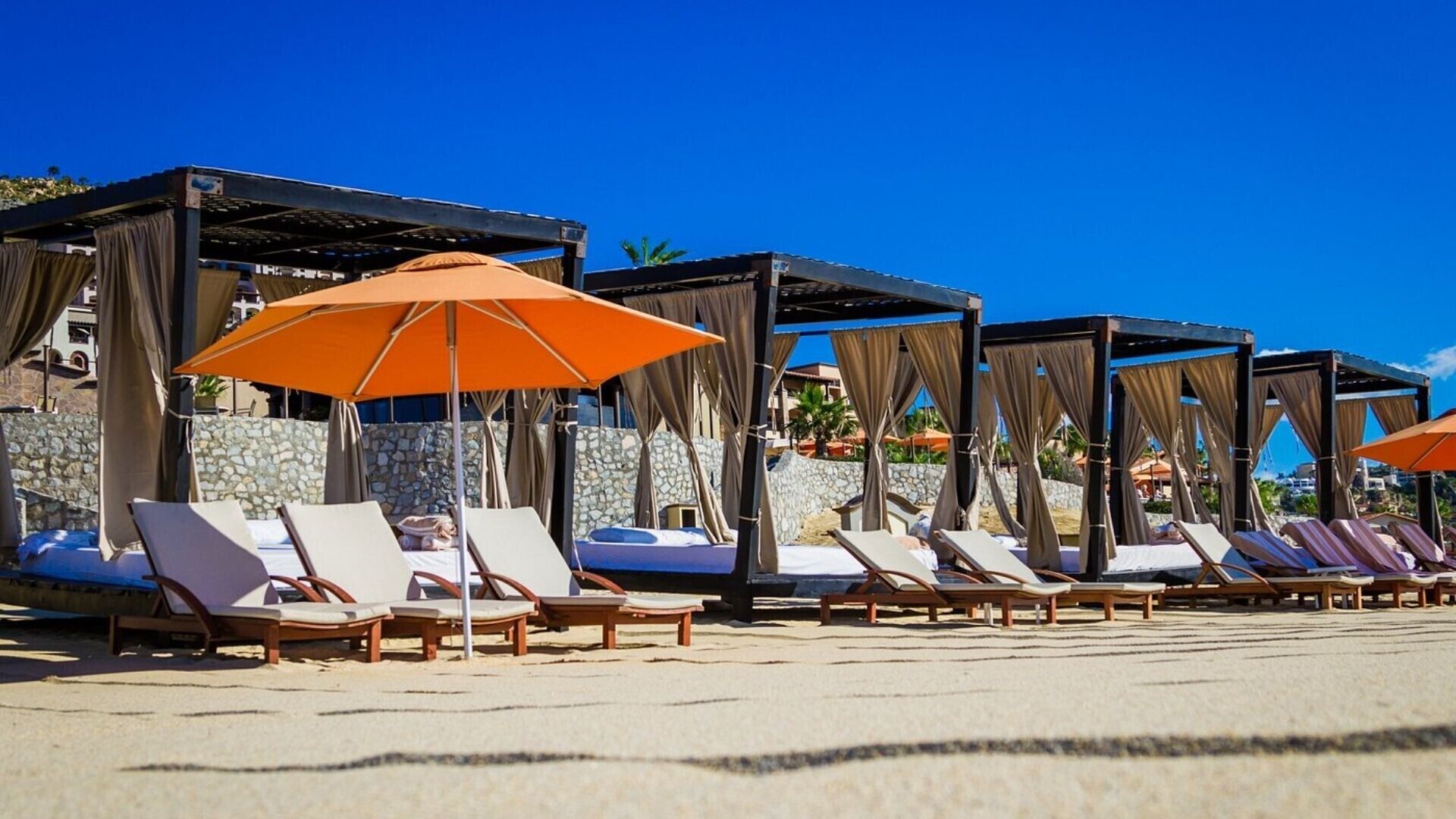 Een resort aan het strand met ligbedden, oranje parasols en stenen muren.