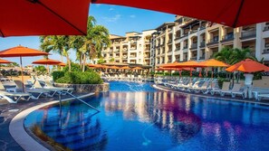 Une piscine entourée de chaises longues et de parasols, elle-même bordée d’un immeuble à plusieurs étages doté de balcons.