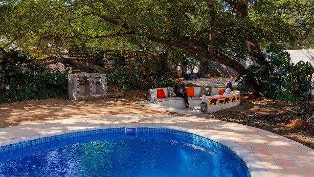 A swimming pool with a tiled edge, surrounded by a garden with trees and a seating area with cushions.