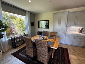 Un coin repas avec une table en bois, des chaises en osier, un miroir au-dessus de la table et une cuisine avec des meubles blancs.