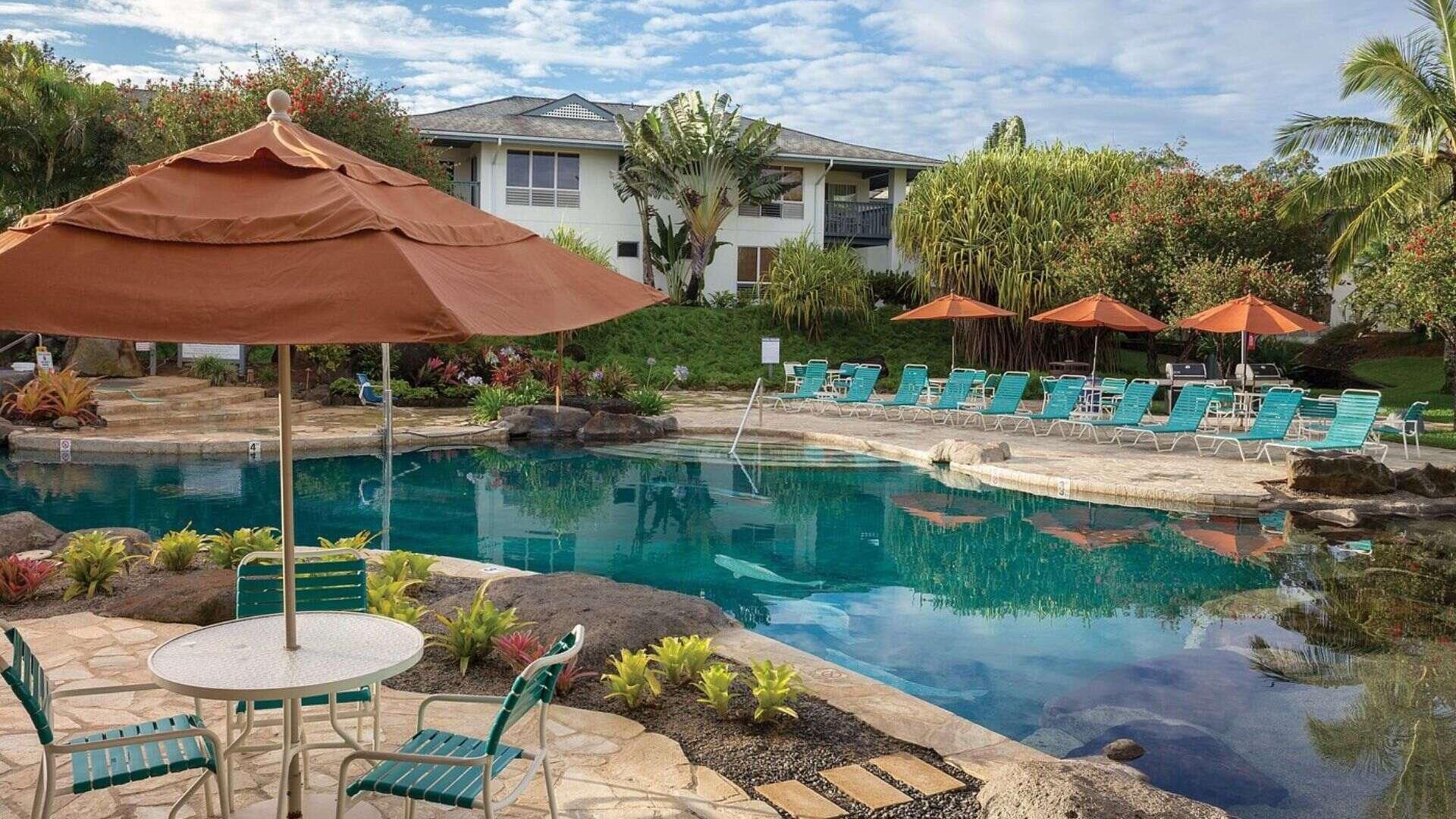 A swimming pool with lounge chairs and umbrellas, surrounded by greenery and a building in the background.
