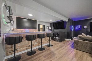 Ein moderner Barbereich mit einer Theke und vier Barhocker, einem flachbildfernseher an der Wand und einem Neonlichtschild.