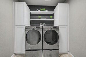 A laundry room with two front-loading washers and dryers, white cabinetry, and a shelf with small potted plants. - Lux Retreat Family-Friendly Near USAFA, Parks (Colorado Springs)