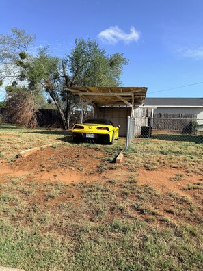 Um carro desportivo amarelo estacionado num quintal cercado, com uma estrutura de madeira ao fundo.