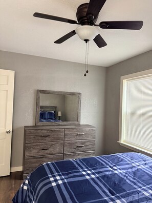 Quarto com cama, penteadeira, ventoinha no teto e janela com persianas.