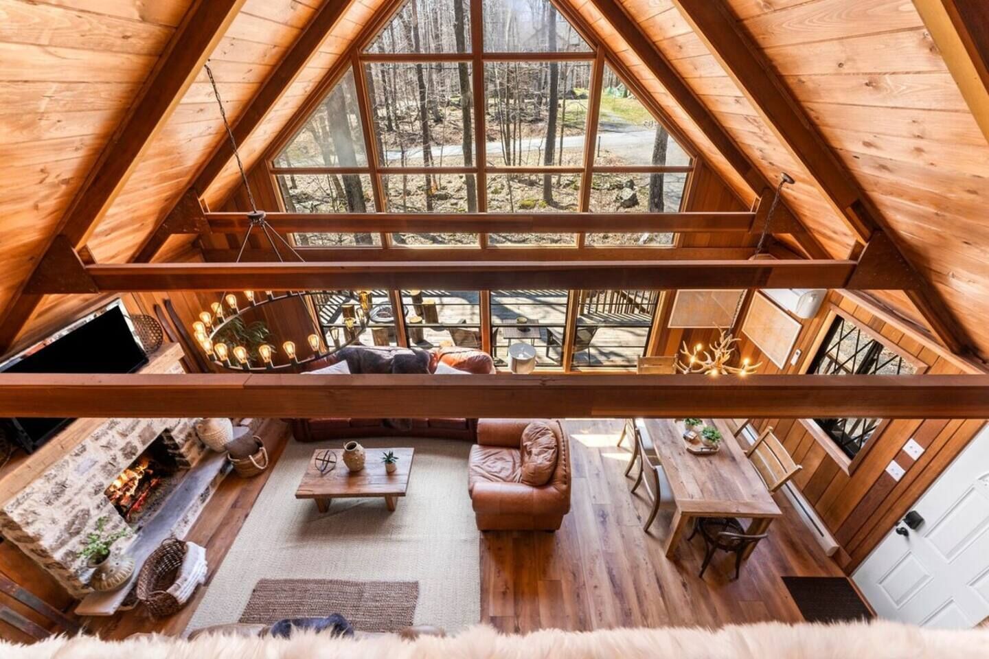 A wooden interior with a loft bedroom, a fireplace, and a dining area with a view of the forest.