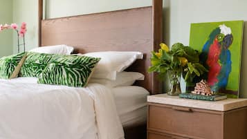 A neatly made bed with white linens and green patterned pillows, a wooden nightstand with a book and a vase of flowers, and a colorful painting of a parrot.