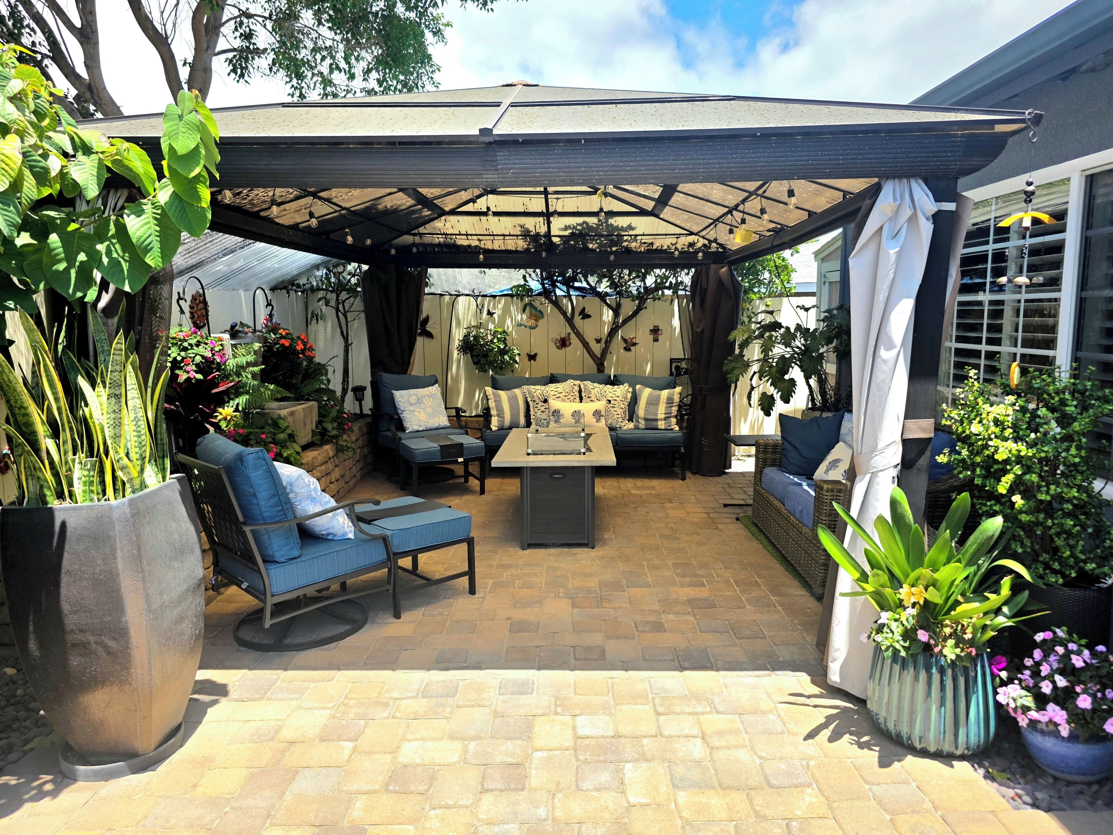 Un patio al aire libre con zona de estar cubierta, que incluye un sofá y sillones, rodeado de plantas en macetas y un pérgola.