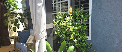 A covered outdoor seating area with a wicker sofa and a potted plant with yellow flowers.
