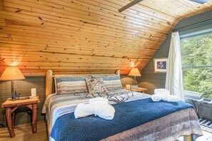 Un dormitorio acogedor con techo inclinado, cama con colcha azul estampada, dos lámparas de noche y ventana con vistas a la vegetación.