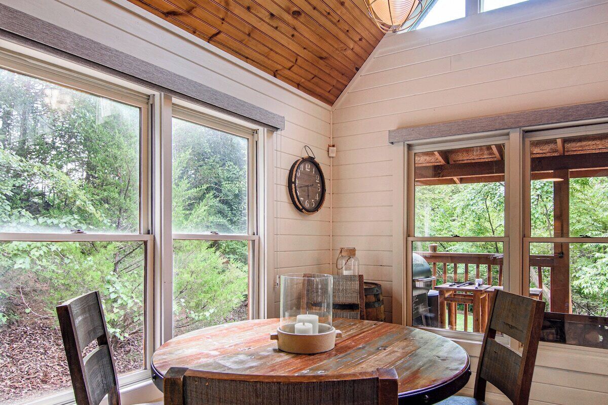 A wooden-paneled room with a round table, chairs, a clock, and large windows offering a view of greenery.