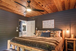 A bedroom with a wooden ceiling, a bed with patterned bedding, a nightstand with a lamp, and a wall sign.