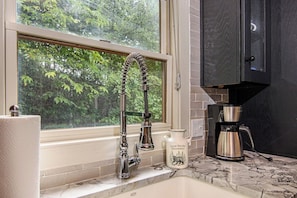 A kitchen with a stainless steel faucet, a coffee maker, and a window with a view of trees.