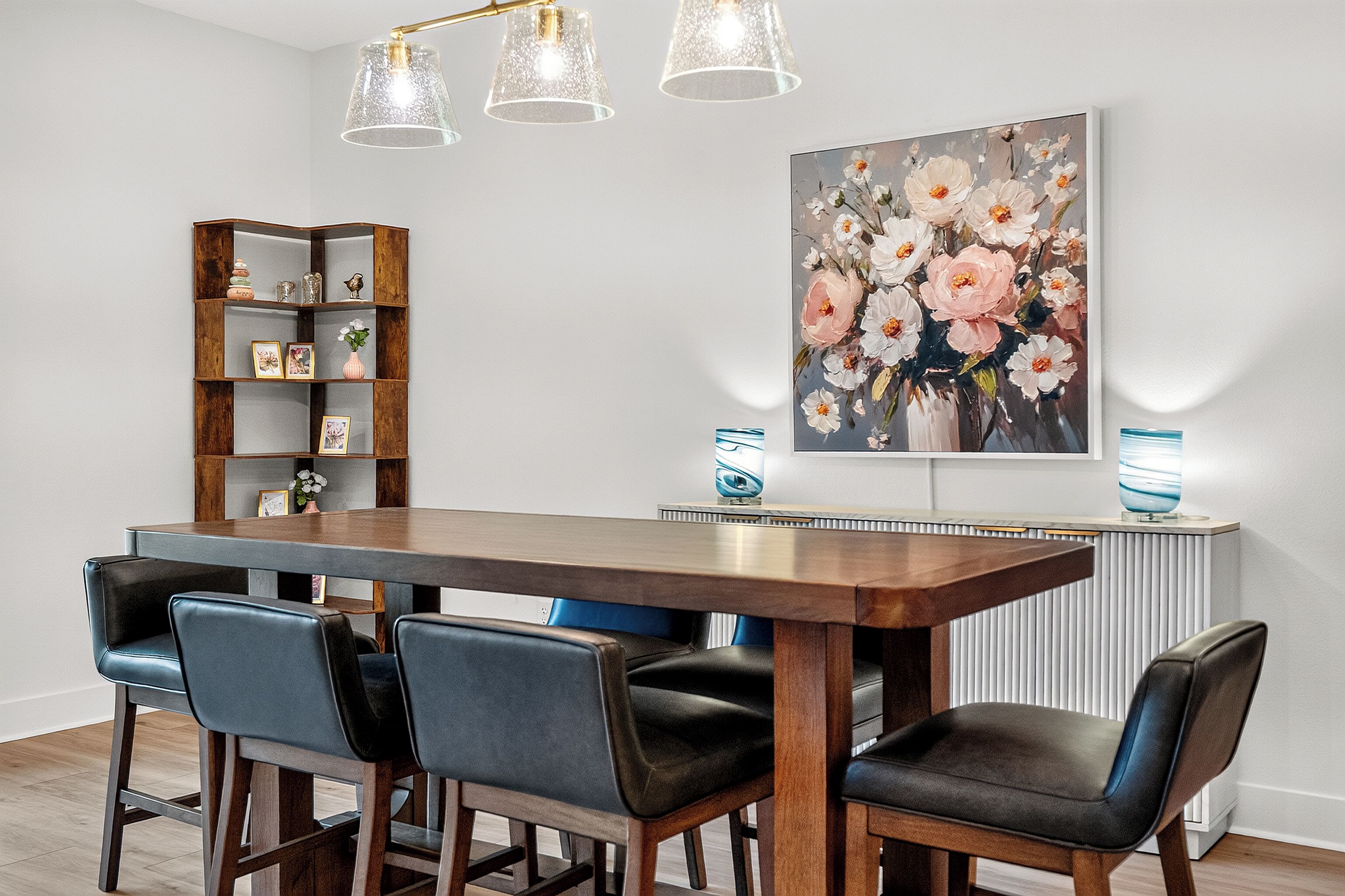 A dining area with a wooden table, four chairs, a bookshelf, and a floral painting on the wall.