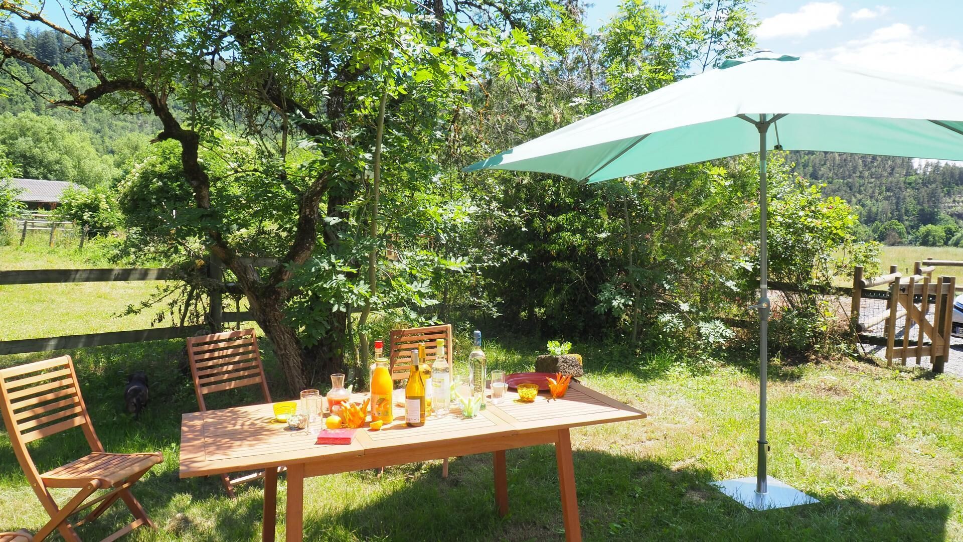 An outdoor dining area with a table set for two, surrounded by greenery and a large umbrella providing shade.