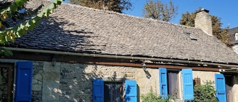 Un edificio de piedra con techo de tejas y contraventanas azules, rodeado de follaje.