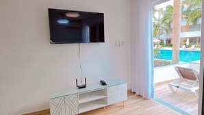 A modern room with a flat-screen TV mounted on the wall, a glass console table, and a view of a pool and palm trees through a large window. - COSTA DE LUXE (Bayahíbe)
