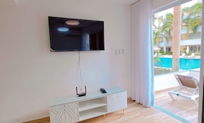 Ein modernes Zimmer mit einem wandmontierten Flachbildfernseher, einem Glastischchen und Blick auf einen Pool und Palmen durch ein großes Fenster.