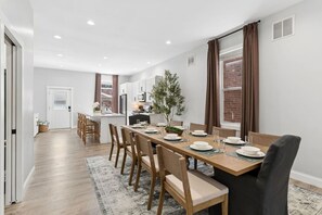 A modern dining area with a long table set for six, wooden chairs, and a kitchen in the background.