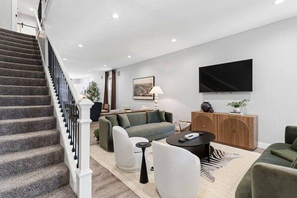 A modern living room with a staircase, a flat-screen TV, a sofa, a coffee table, and a sideboard.