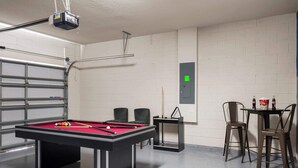 Ein Billardtisch mit roter Spielbahn, zwei schwarze Stühle, ein Tisch mit Flaschen und ein an der Decke befestigter Projektor.