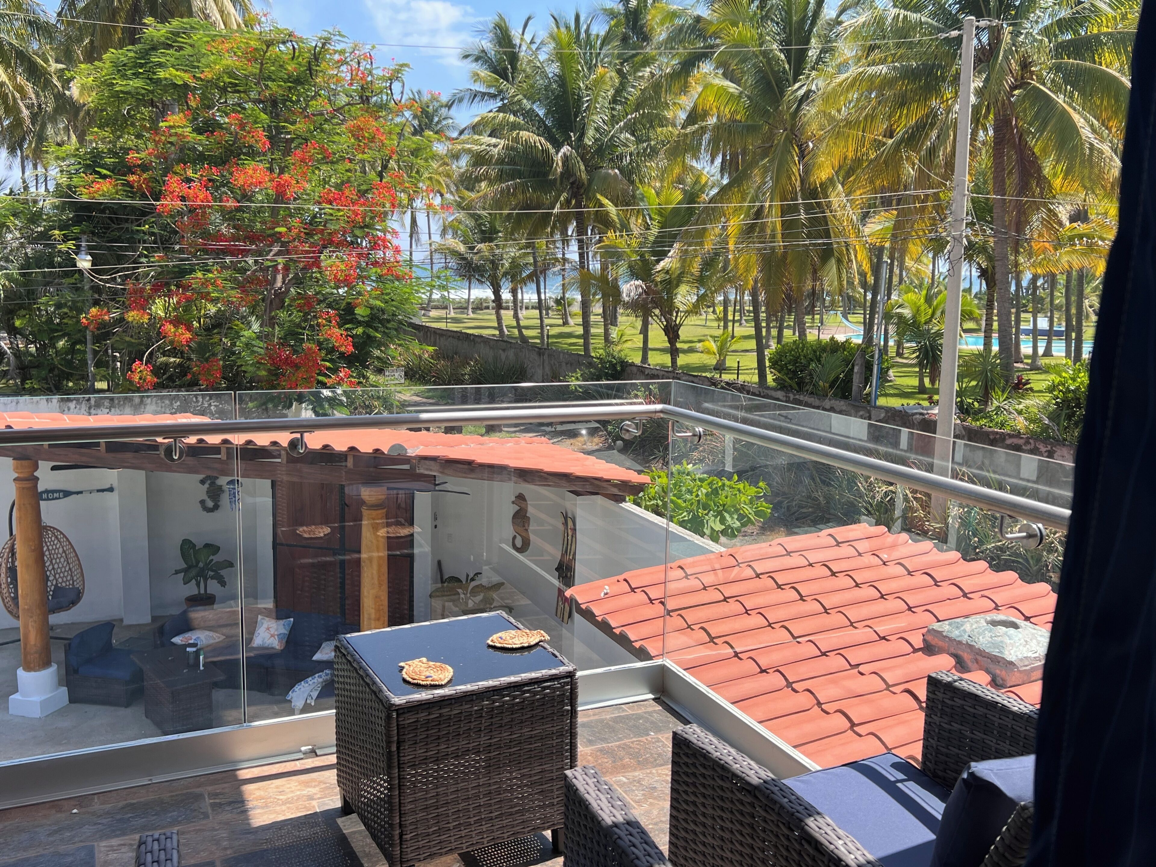 Uma varanda com móveis de vime, guarda-corpo de vidro e vista para uma área ajardinada com palmeiras e uma piscina.