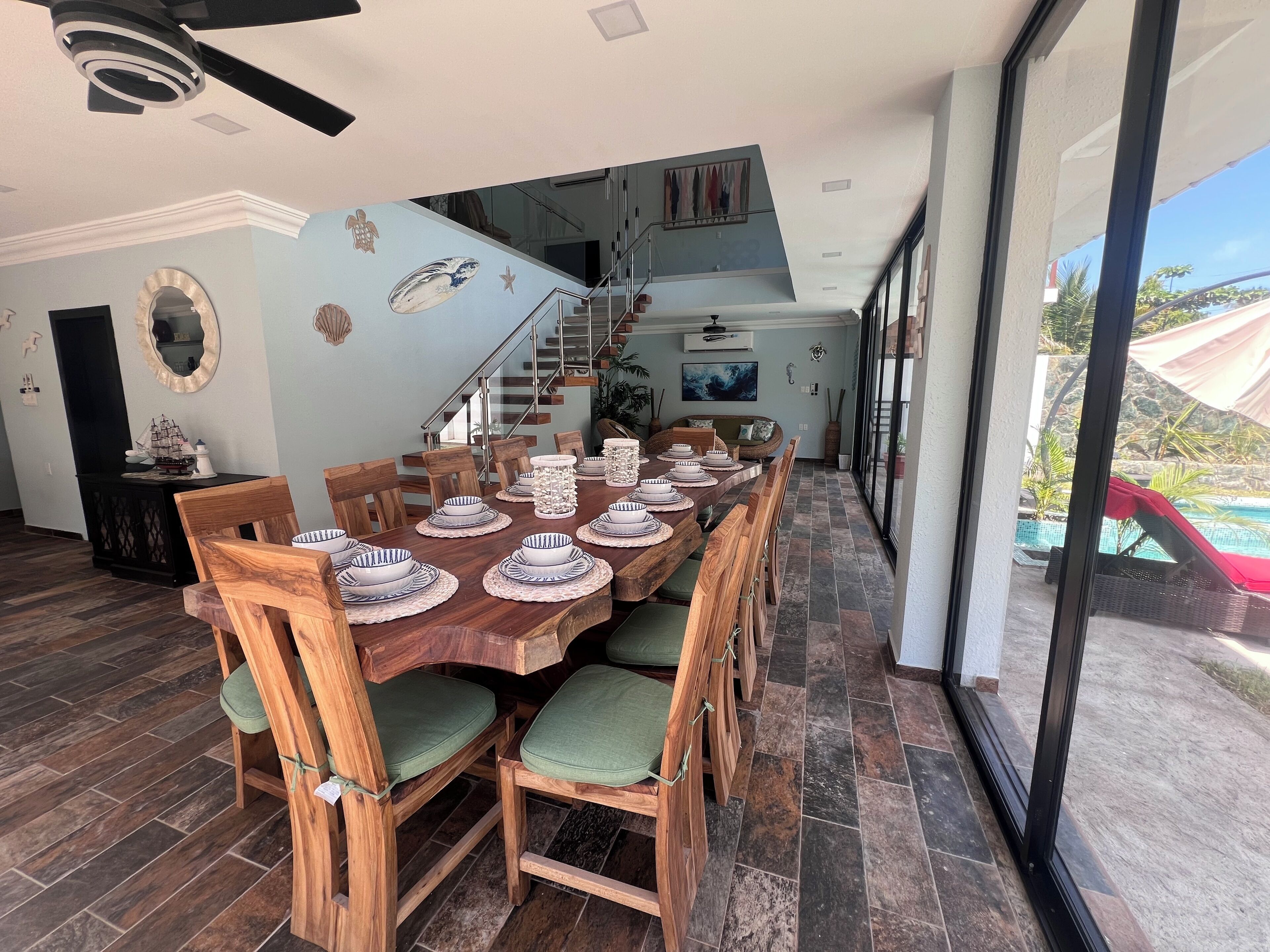 Une vaste salle à manger avec une longue table en bois dressée pour le repas, un escalier menant à un niveau supérieur et une vue sur une piscine à l’extérieur.