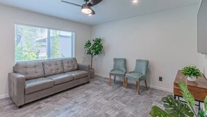 Un salon avec un canapé gris, deux fauteuils à motifs, un ventilateur de plafond et une grande fenêtre avec des stores.